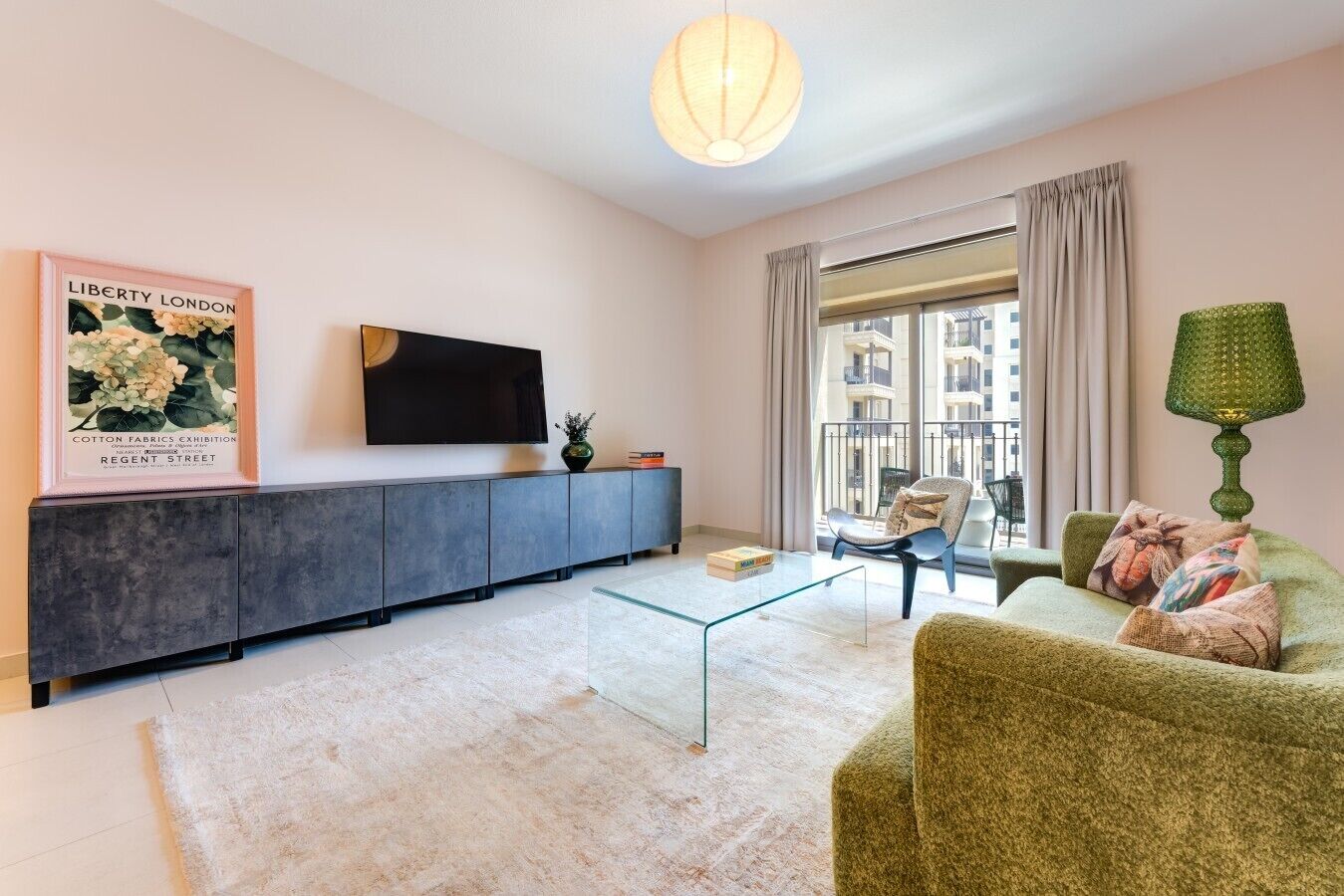 Un salon moderne avec un canapé gris, une table basse en verre, un téléviseur à écran plat fixé au mur et une grande fenêtre avec des rideaux.