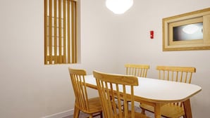 Un comedor con una mesa blanca y cuatro sillas de madera, una ventana con persianas verticales, una lámpara colgante y un espejo enmarcado en la pared.