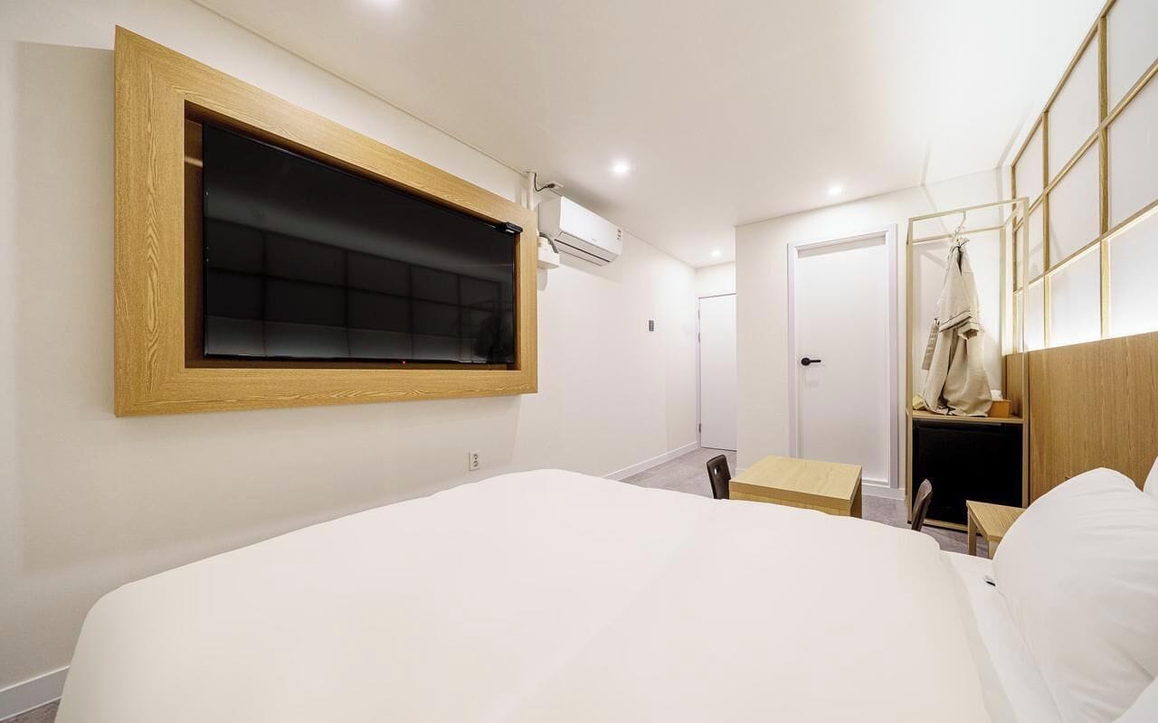Habitación de hotel con una cama grande, ventana con marco de madera, una mesita pequeña, una silla y un armario con espejo.