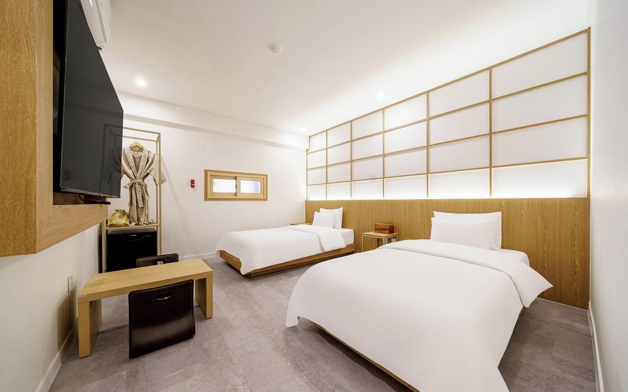 A hotel room with two beds, a TV, a wooden desk, and a bench.