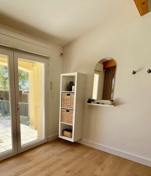 A room with a sliding glass door leading to an outdoor area, a white shelving unit with baskets, and a mirror on the wall. - BY THE WATER'S EDGE (Frontignan)