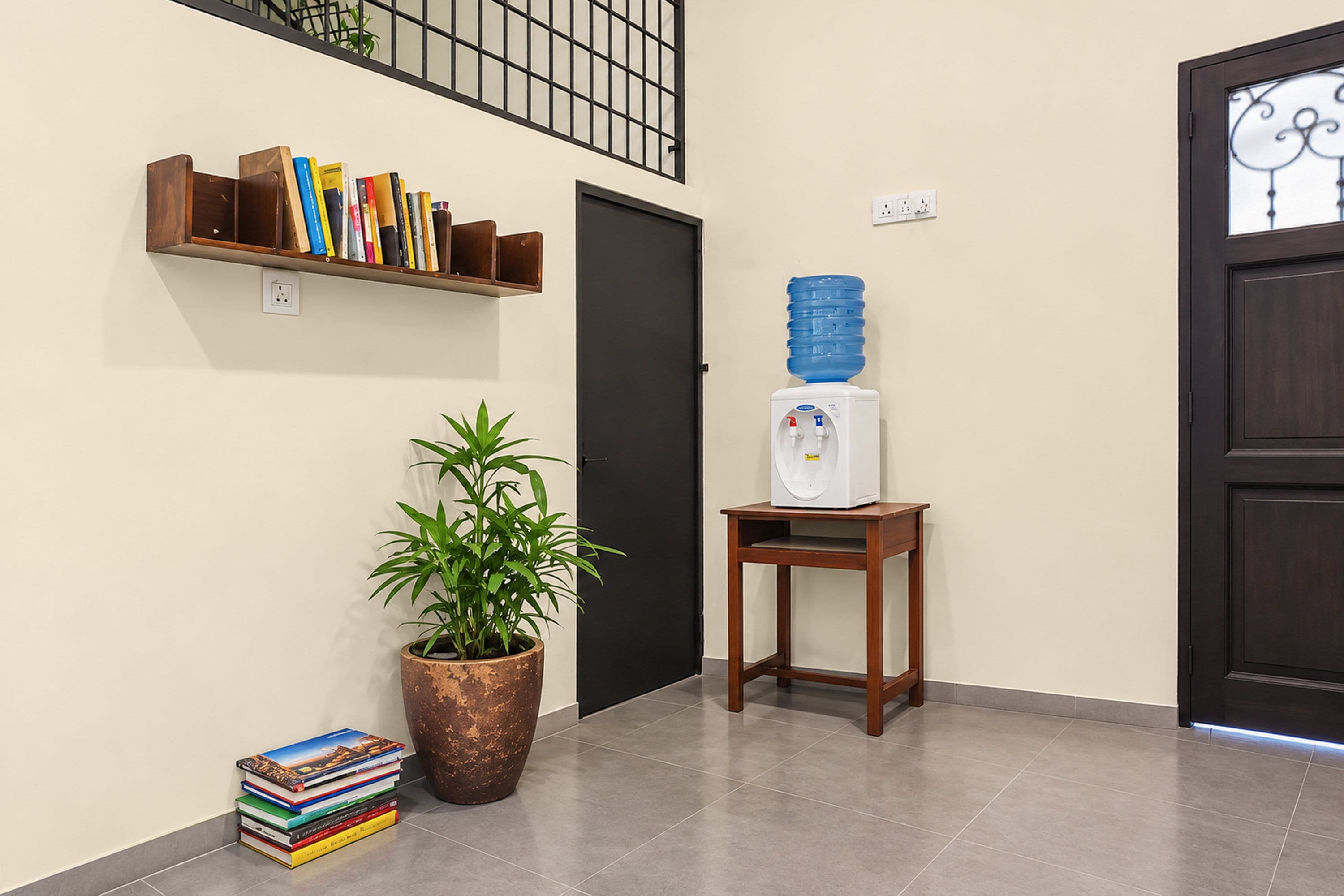 Corredor com estante, bebedouro, planta em vaso e pilha de livros.
