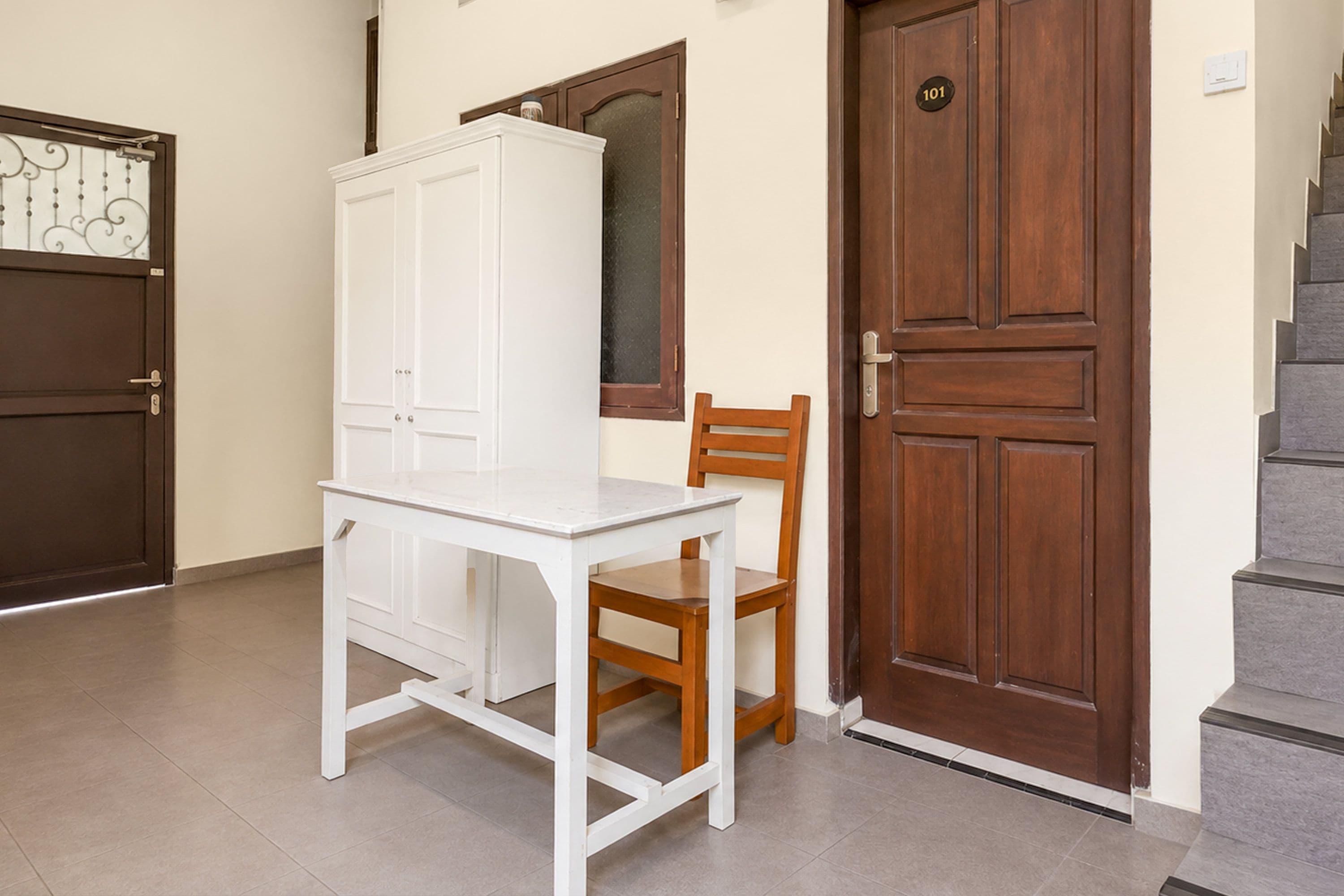 A room with a white desk, a wooden chair, a white wardrobe, and a brown door with a number plate.