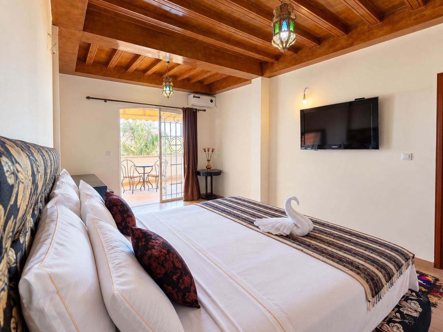 Habitación de hotel con cama, televisión, balcón con mesa y sillas, y un cisne decorativo sobre la cama.