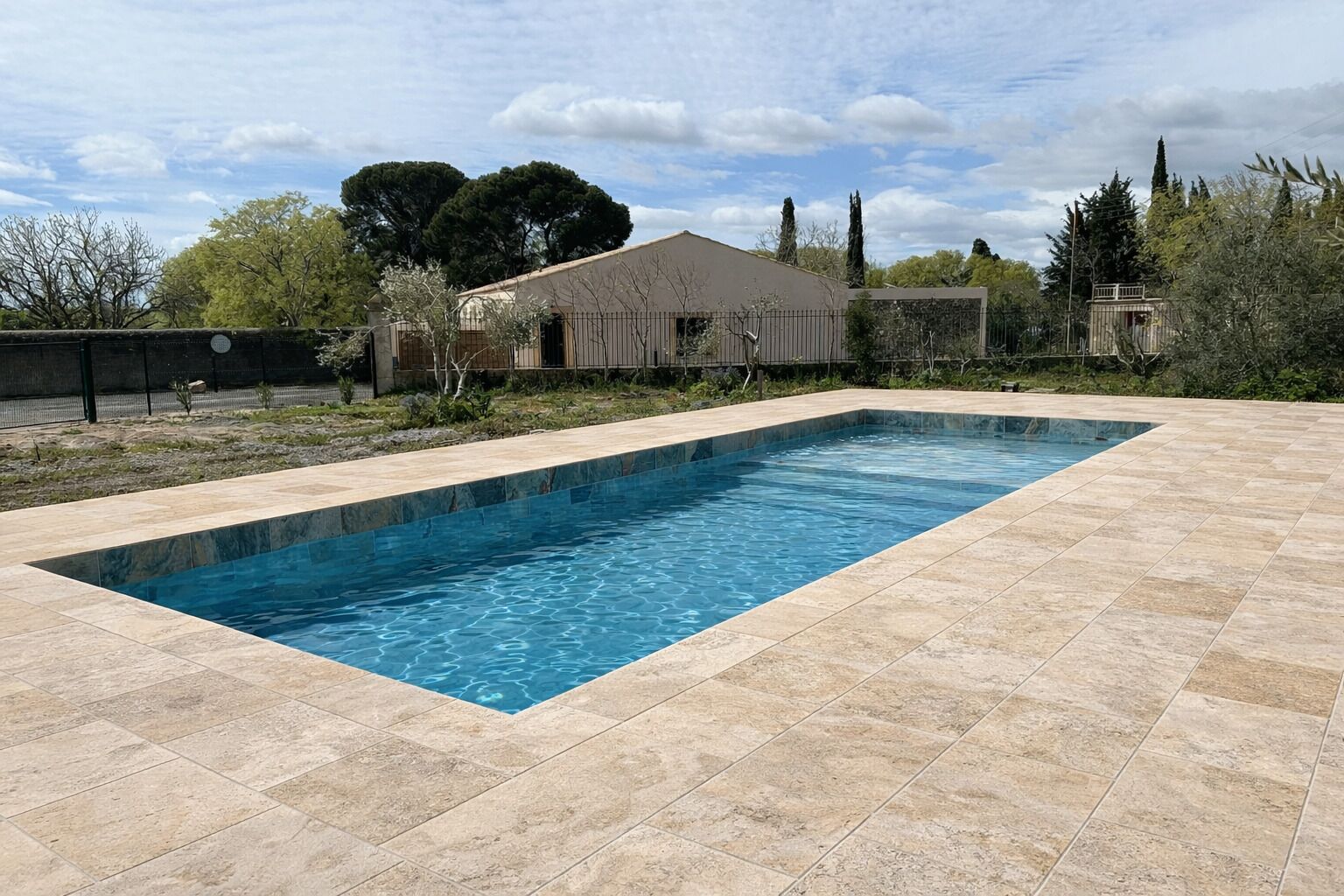 A rectangular swimming pool with a tiled deck, surrounded by a fence and trees.