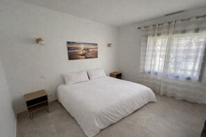 A bedroom with a bed, a nightstand, a wall-mounted lamp, a painting, and a window with sheer curtains. - Spacious 4-bedroom house in Florensac with AC (florensac)