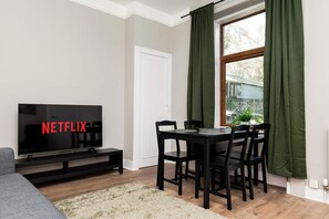 A living room with a television displaying 'NETFLIX,' a dining table with chairs, a sofa, and a window with curtains. - Spacious Inverurie Stay -Nr Train Station -Parking (Aberdeenshire)