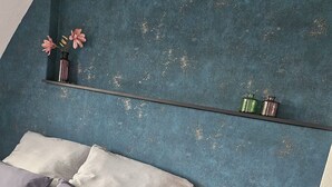 Een slaapkamer met een bed, kussens, een nachtkastje, een klein tafeltje en een plank met decoratieve objecten.