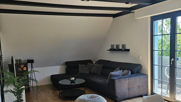 Een moderne woonkamer met een donkere bank, een rond hocker en een kleine tafel. Een groot raam zorgt voor natuurlijk licht en uitzicht.