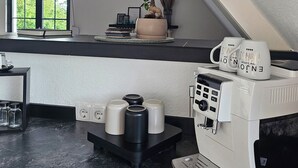 Een keuken met een koffieapparaat, een raam met uitzicht op groen en een plant op het aanrecht.