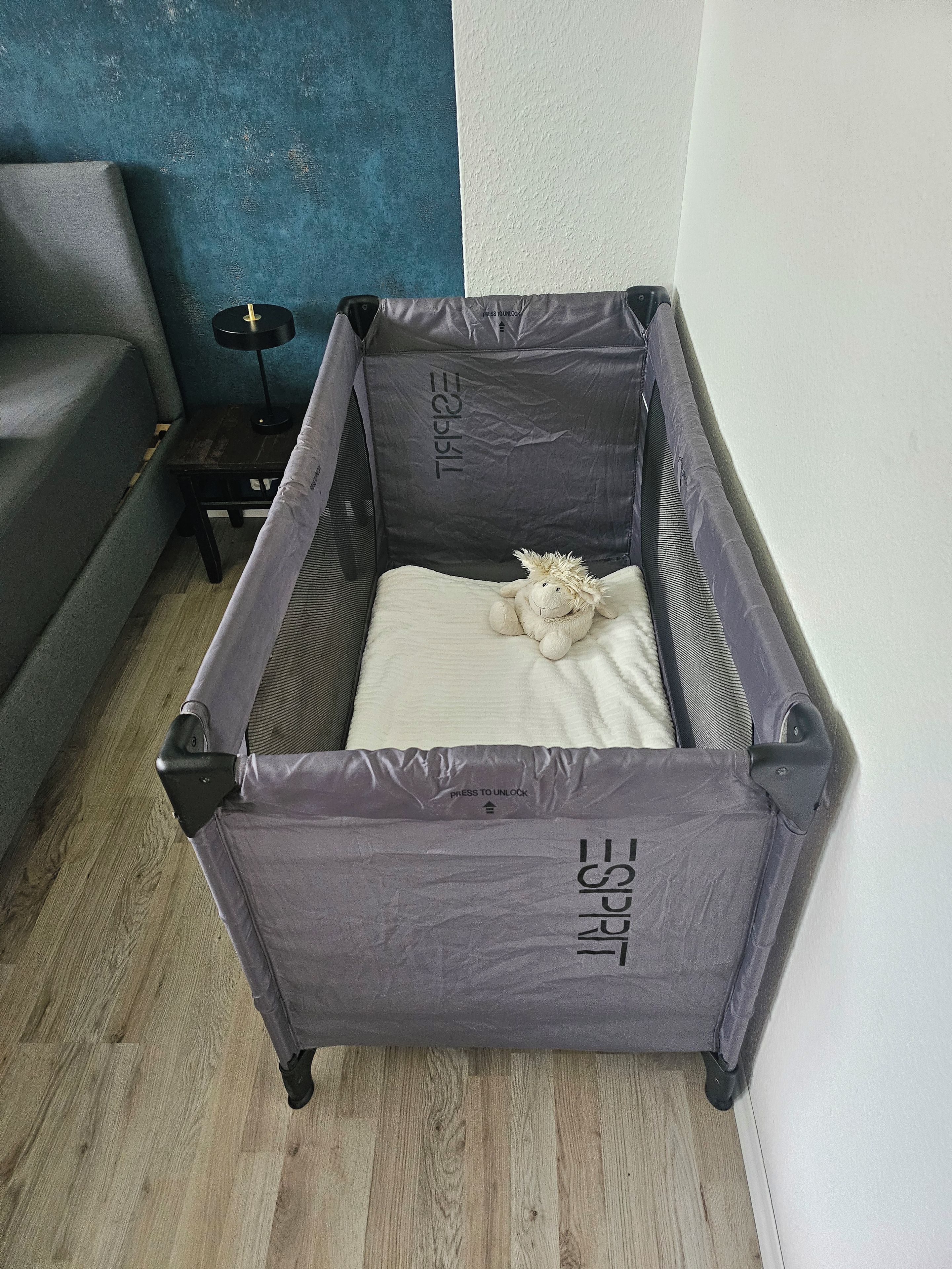 Ein kleines, graues, zusammenklappbares Babybett mit einer weißen Matratze und einem Stofftier im Inneren, vor einer blauen Wand und einem grauen Sofa platziert.