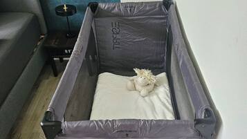 Ein kleines, graues, zusammenklappbares Babybett mit einer weißen Matratze und einem Stofftier im Inneren, vor einer blauen Wand und einem grauen Sofa platziert.