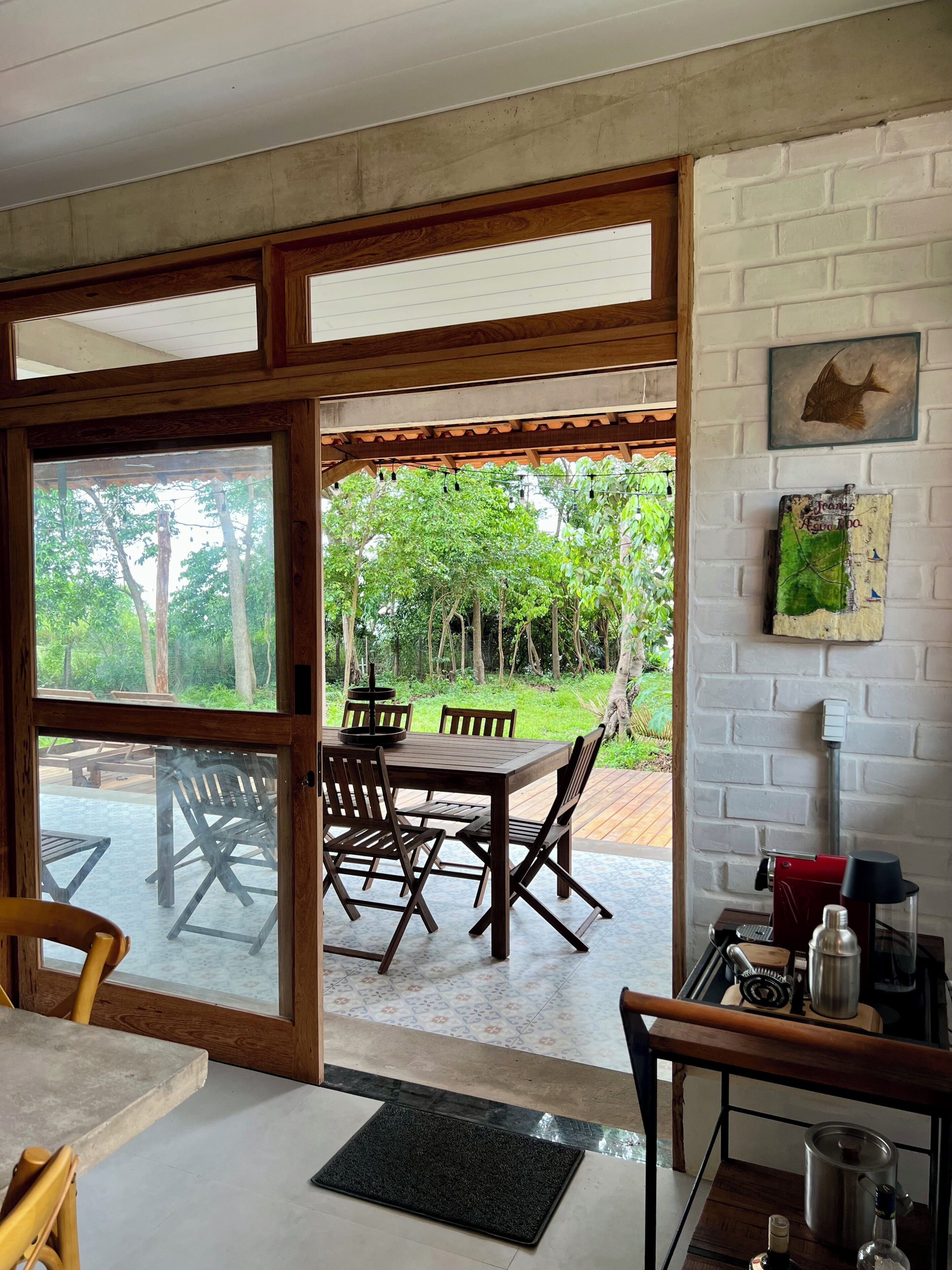A room with a wooden dining table and chairs, a coffee machine, and a view of an outdoor patio with wooden tables and chairs.