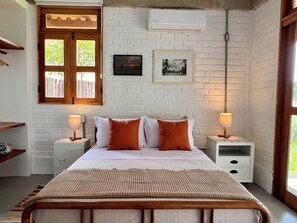 Une chambre à coucher comprenant un lit, deux tables de chevet, une fenêtre, un climatiseur fixé au mur et un tableau encadré.