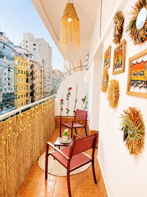 A balcony with a view of city buildings, a woven light fixture, a wooden chair, a small table, and decorative wall art. - Nejma House with a balcony—5 minutes from the beach. (Tangier)