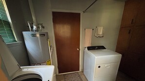 Ett tvättrum med tvättmaskin och torktumlare, en varmvattenberedare och en dörr som leder till ett annat rum.