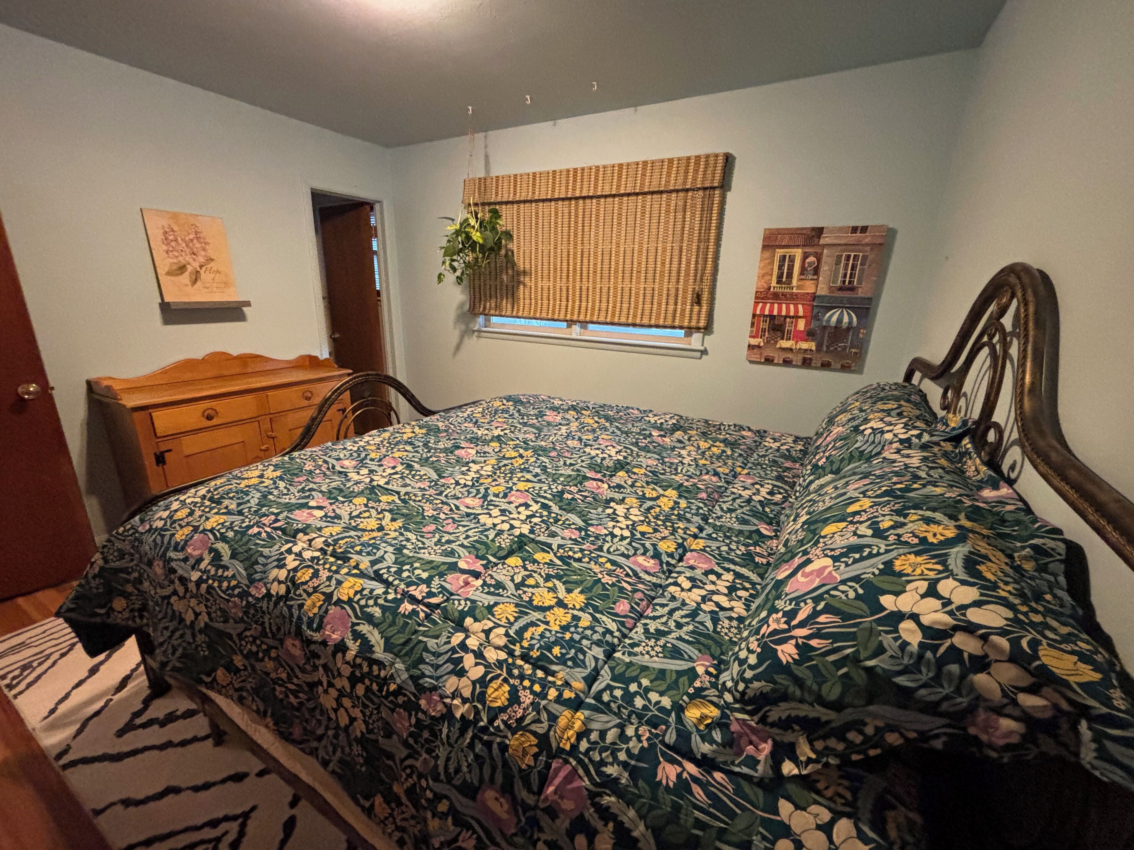 Une chambre à coucher avec une couvre-lit à motifs floraux, une commode en bois, une fenêtre avec une plante et un mur orné d’un tableau.