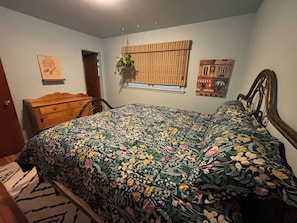 Un dormitorio con cubrecama floral, un aparador de madera, una ventana con planta y un cuadro en la pared.
