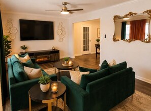 A living room with a green sofa set, a flat-screen TV, a fireplace, and a ceiling fan. - Emerald Escape close to Airport,NRG,Medical center & ZOO With PS5 &Foosball (Houston)