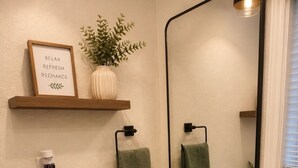 Et badeværelse med en hvid vask, en sort vandhane, et spejl, et håndklædestativ, et indrammet skilt, en potteplante og en toiletpapirholder.
