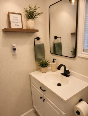 A bathroom with a white sink, black faucet, a mirror, a towel rack, a framed sign, a potted plant, and a toilet paper holder. - Emerald Escape close to Airport,NRG,Medical center & ZOO With PS5 &Foosball (Houston)