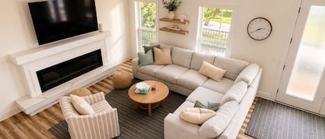A modern living room with a sectional sofa, a television mounted above a fireplace, and a large window providing natural light.
