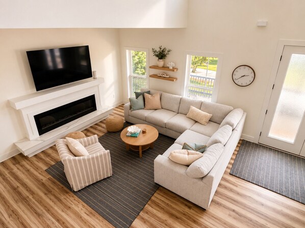 A modern living room with a sectional sofa, a television mounted above a fireplace, and a large window providing natural light.