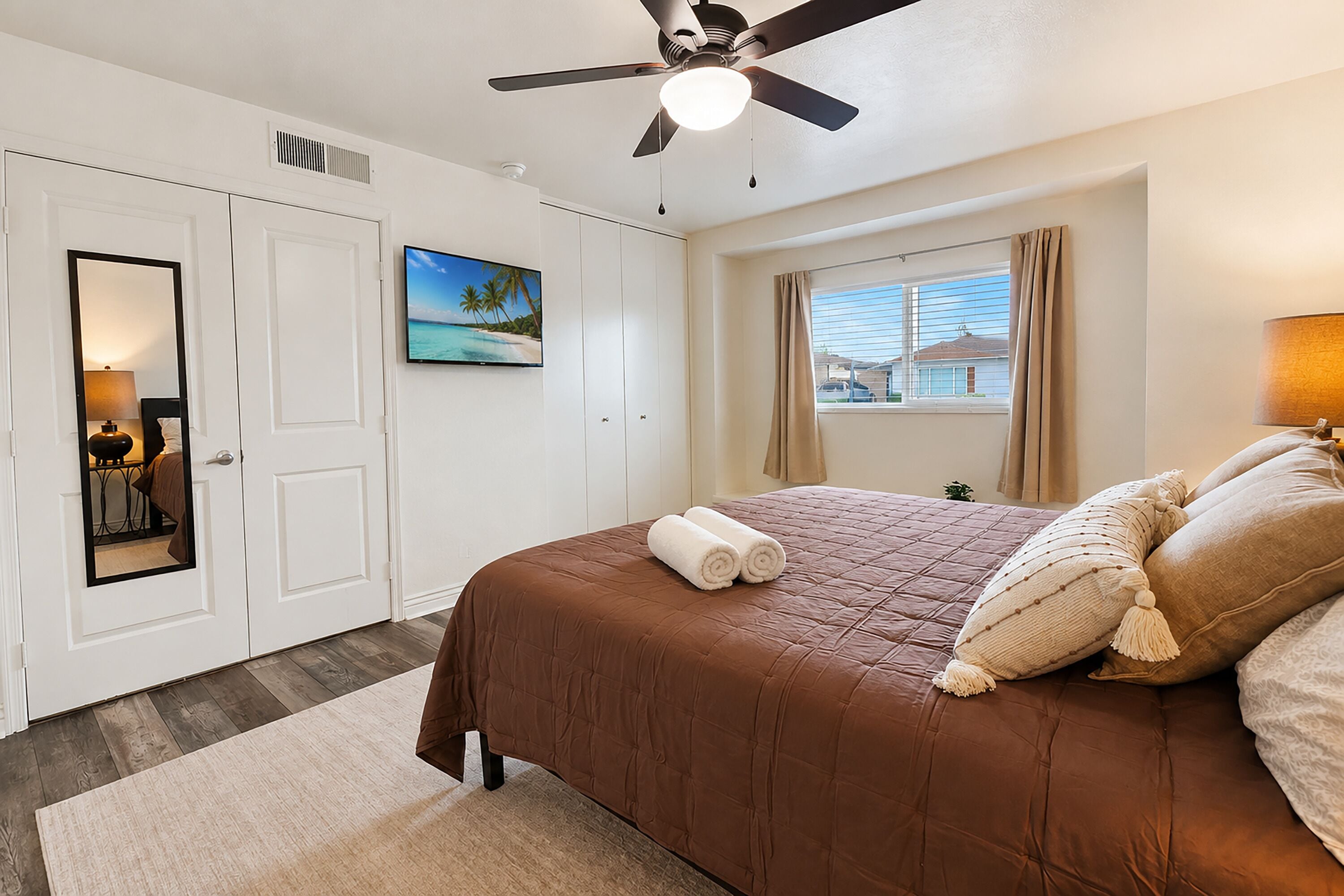 A bedroom with a bed, a ceiling fan, a window with curtains, a door, and a wall-mounted TV.