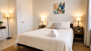 A neatly made bed with rolled towels, two bedside tables with lamps, a door, and a wall art piece. - Aloha Scottsdale, Poolside Old Town Retreat (Scottsdale)