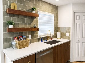Eine Küche mit weißer Arbeitsplatte, Spüle aus Edelstahl, Holzschränken und einer Rückwand aus grauen Fliesen.