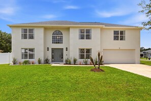 A two-story house with a front garage, a central entrance with a door, and a small front yard with a tree. - 7BR Heated Pool | Game Room + Private Suite (Palm Coast)