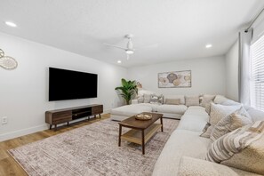 A modern living room with a flat-screen TV, a wooden entertainment unit, a large sofa, a coffee table, and a decorative wall piece. - 7BR Heated Pool | Game Room + Private Suite (Palm Coast)