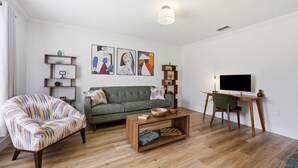 Uma sala de estar com um sofá verde, uma poltrona estampada, uma mesa de centro de madeira e uma televisão sobre um móvel.
