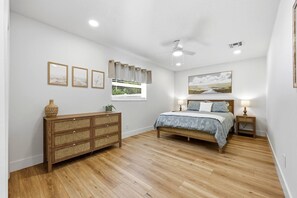 Ein Schlafzimmer mit einem hölzernen Schminktisch, einem Bett mit blau-weißer Bettdecke, einem Deckenventilator und einem Fenster mit Blick auf Grünfläche.