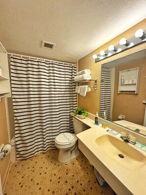A bathroom with a striped shower curtain, a toilet, a sink with a mirror above it, and a shelf with toiletries. - NEW Amazing Ocean Front Studio-Up to 4 Guests! (Daytona Beach Shores)