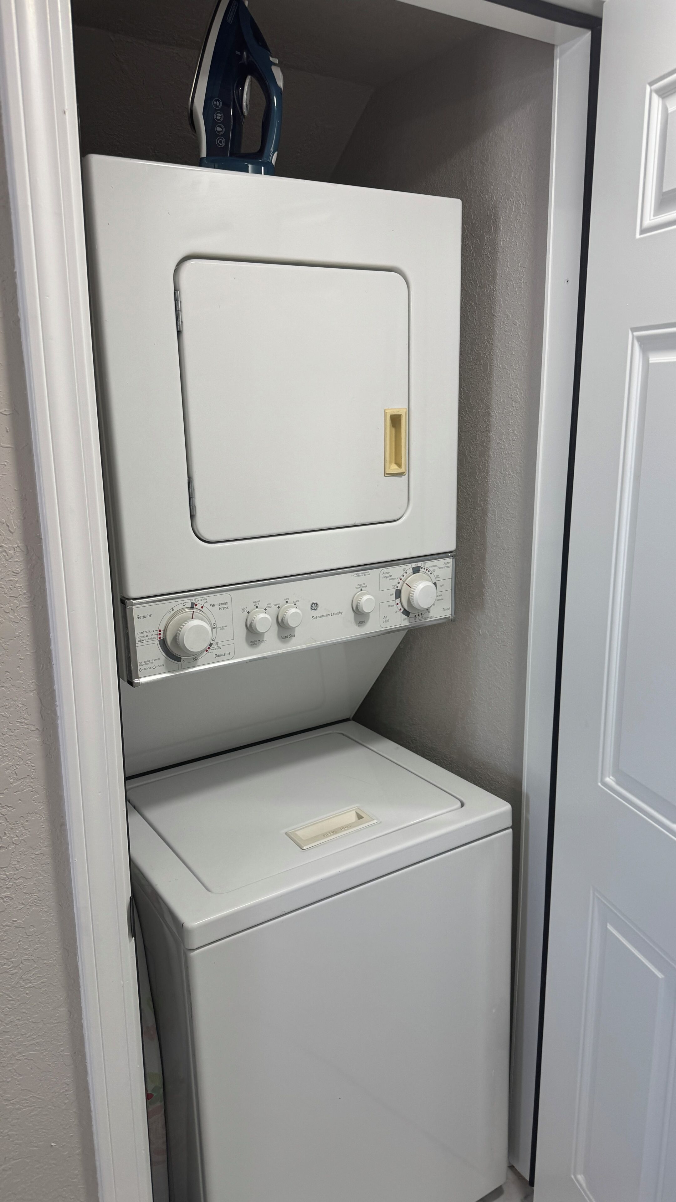 A compact laundry room with a front-loading washer and dryer, a control panel with various settings, and a small shelf above the dryer.