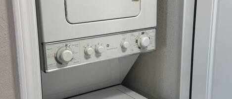 A compact laundry room with a front-loading washer and dryer, a control panel with various settings, and a small shelf above the dryer.