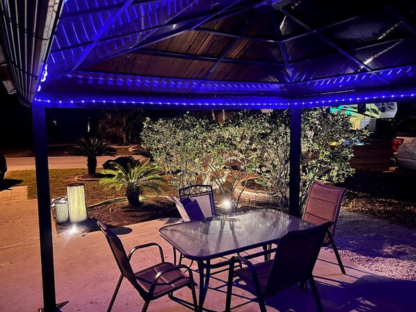 Un patio exterior con una mesa y sillas de vidrio, rodeado de plantas y una cerca de madera.