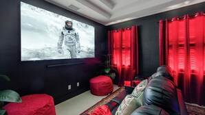 Una habitación con una televisión de pantalla plana grande montada en la pared, un sofá rojo, un reposapiés rojo y una alfombra estampada.