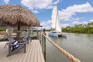 Eine Holzterrasse mit Liegestühlen und einem strohgedeckten Bauwerk mit Blick auf ruhiges Wasser und Segelboote.