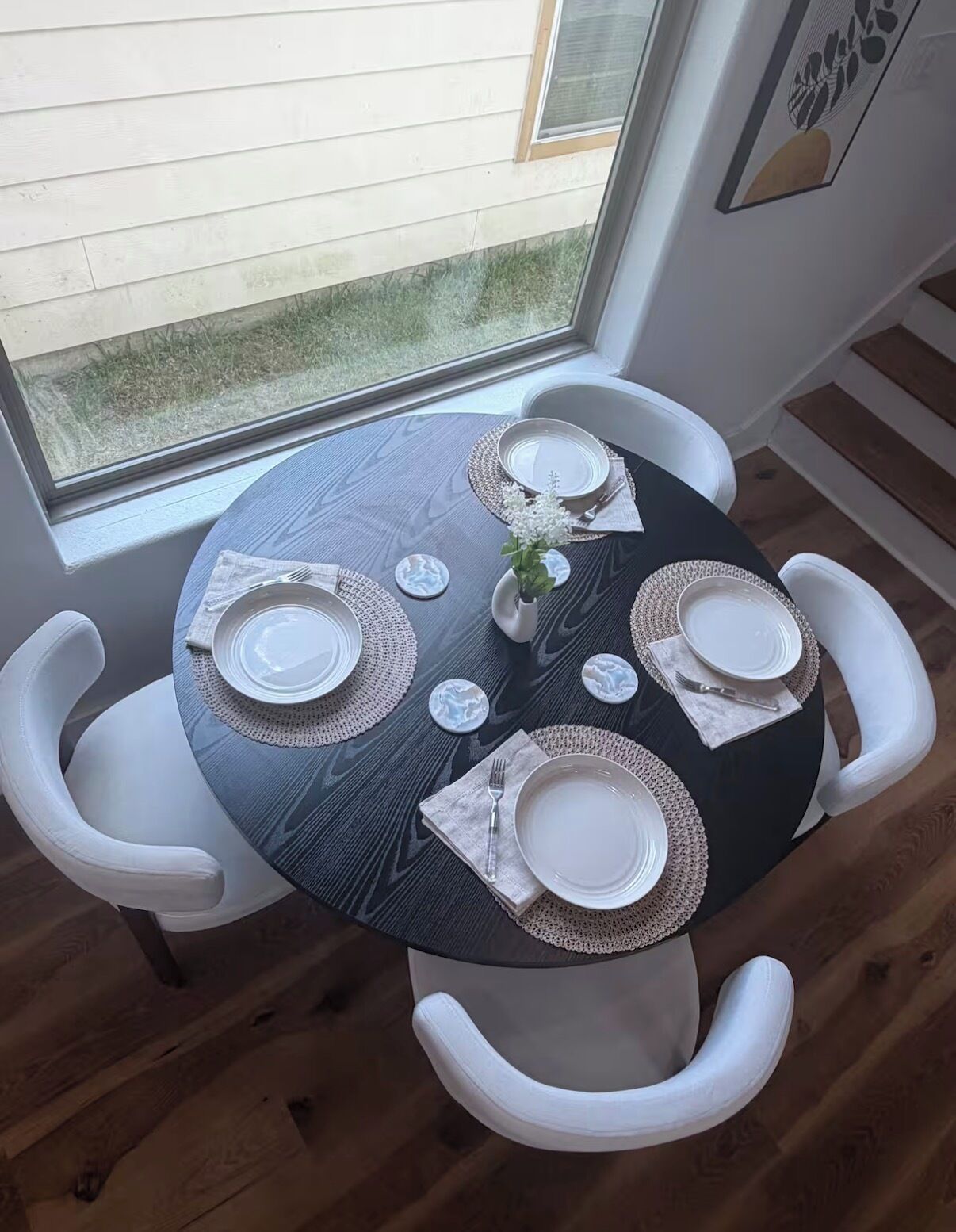 A round dining table with white chairs, set with plates, cutlery, and a small vase with flowers.