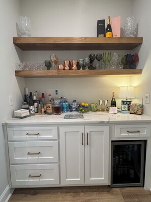 Une cuisine avec une étagère en bois où sont rangés divers objets en verre et des bouteilles, un plan de travail avec des ustensiles de cuisine et un réfrigérateur.