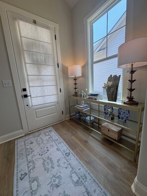 Un couloir avec une porte blanche, une console en verre ornée d’objets décoratifs, un tapis à motifs et une fenêtre avec des stores.