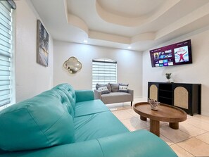 A living room with a teal sofa, a grey armchair, a wooden coffee table, and a flat-screen TV mounted on the wall.