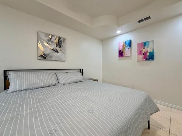 A bedroom with a bed, two wall-mounted paintings, and a ceiling vent.
