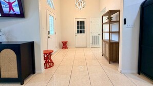 A hallway with a television displaying a program, a door with a window, a red chair, a white door, and a bookshelf. - Casa Pera - Minutes to SpaceX & Port - Spacious! (Brownsville)