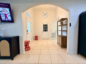 A hallway with a television displaying a program, a door with a window, a red chair, a white door, and a bookshelf. - Casa Pera - Minutes to SpaceX & Port - Spacious! (Brownsville)