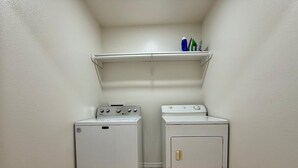A laundry room with two white washing machines and a shelf above them. - Casa Pera - Minutes to SpaceX & Port - Spacious! (Brownsville)
