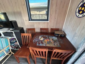 Ein Holzespeisetisch mit einem Puzzle in der Mitte, umgeben von Stühlen, einem Fernseher links und einem Fenster mit Blick auf ein Gebäude.