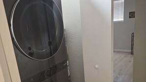 Un lavadero con dos lavadoras y secadoras apiladas, una puerta blanca que da a otra habitación, y una alfombra pequeña en el suelo.